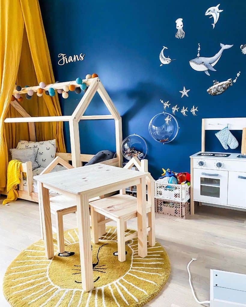 Playful kids' room with a deep blue ocean-themed wall, wooden house bed, yellow canopy, mini kitchen set, and cozy play area