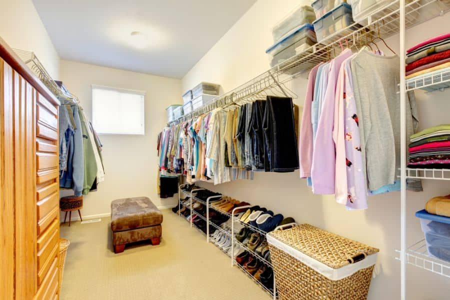 Spacious walk-in closet with clothes hanging, folded sweaters, shoes on shelves, storage boxes, and a brown ottoman on the beige carpet