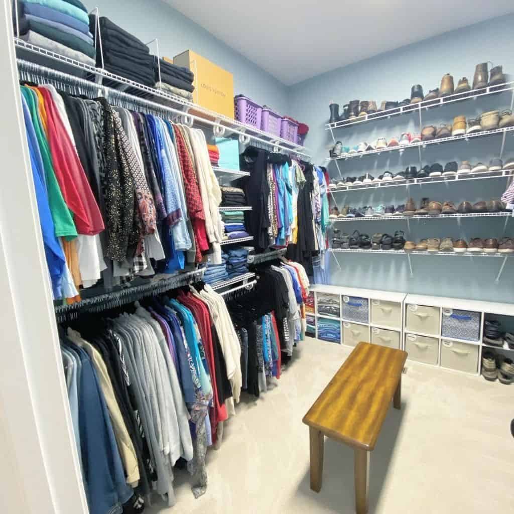 A tidy walk-in closet with clothes on hangers, organized shelves of shoes, and storage bins, featuring a wooden bench in the center