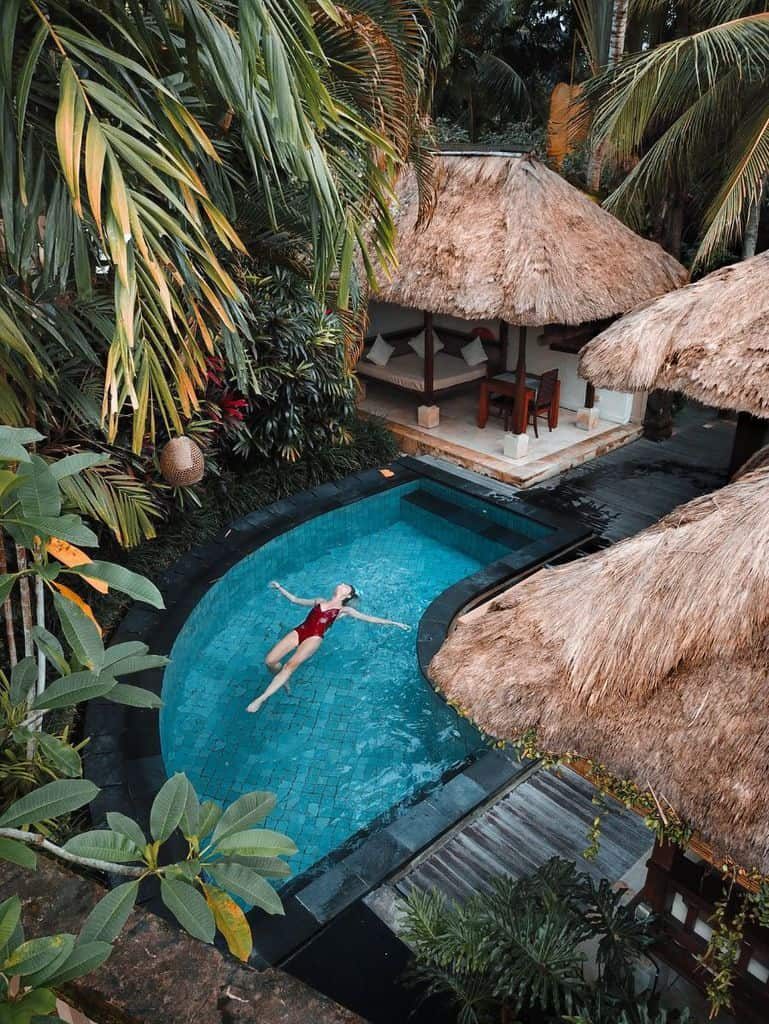 Tropical poolside cabana with thatched roofs, lush greenery, and a woman floating in a serene, curved pool for ultimate relaxation