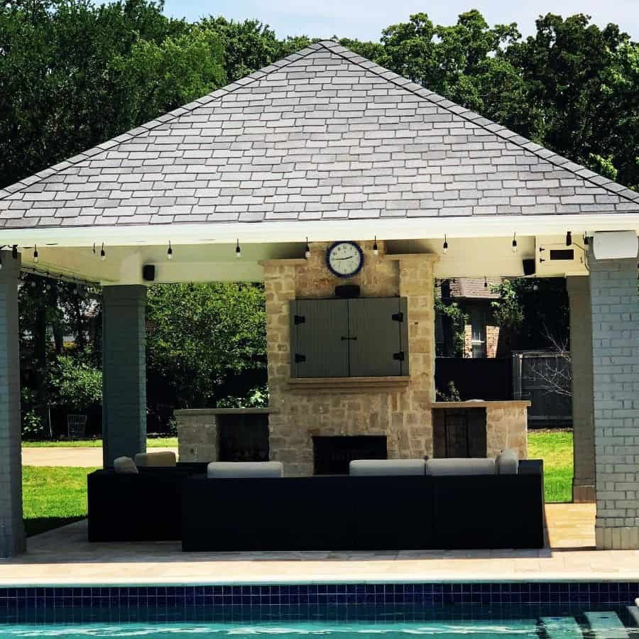 Stylish poolside cabana with a gabled roof, stone fireplace, outdoor kitchen, and cozy seating, perfect for relaxing and entertaining