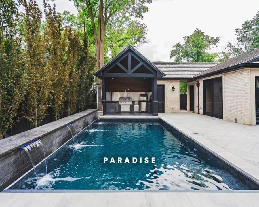 Modern poolside cabana with a sleek outdoor kitchen, black timber beams, and cascading water features, creating a luxurious backyard retreat