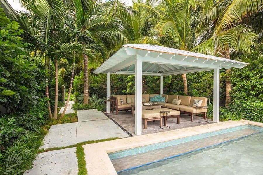 Charming poolside cabana with a white pergola, cozy sectional seating, and lush tropical greenery, creating a serene outdoor lounge space