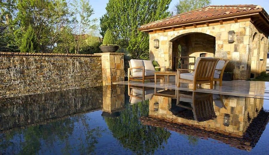 Elegant Mediterranean-style poolside cabana with stone walls, arched entryway, and cozy outdoor seating, reflecting on a serene pool