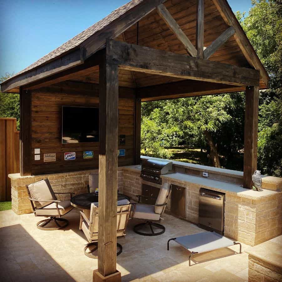 Rustic outdoor cabana with exposed wood beams, a built-in kitchen, TV, and cozy seating, creating a stylish backyard entertainment space