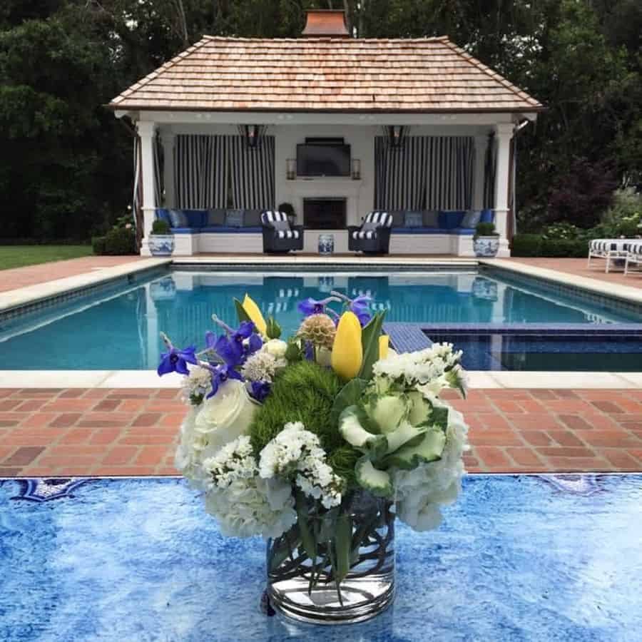 Elegant poolside cabana with a shingled roof, striped curtains, cozy seating, and a fireplace, framed by a vase of fresh flowers