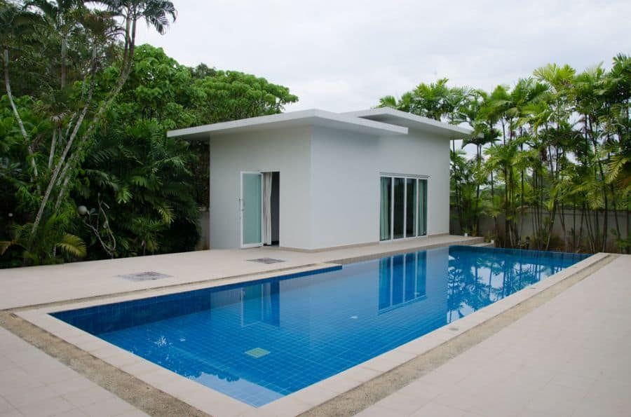 Modern minimalist pool house with a sleek white exterior, large glass doors, and a serene blue-tiled pool, surrounded by lush greenery