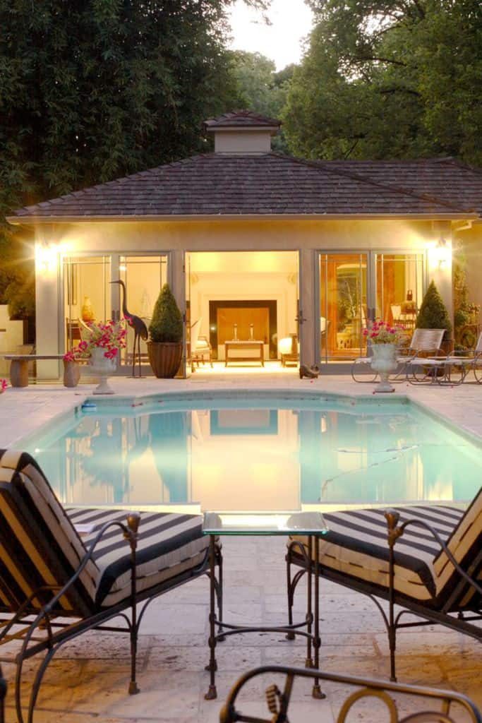 Elegant pool house with warm lighting, large glass doors, and a cozy fireplace, creating a luxurious and inviting backyard retreat