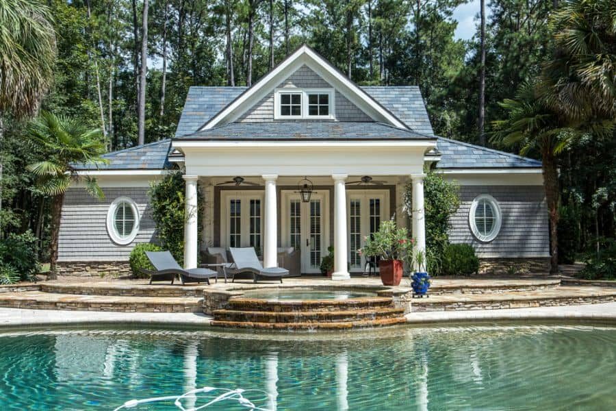 Classic pool house with a gabled roof, elegant columns, and lush greenery, overlooking a serene pool with stone steps and lounge chairs