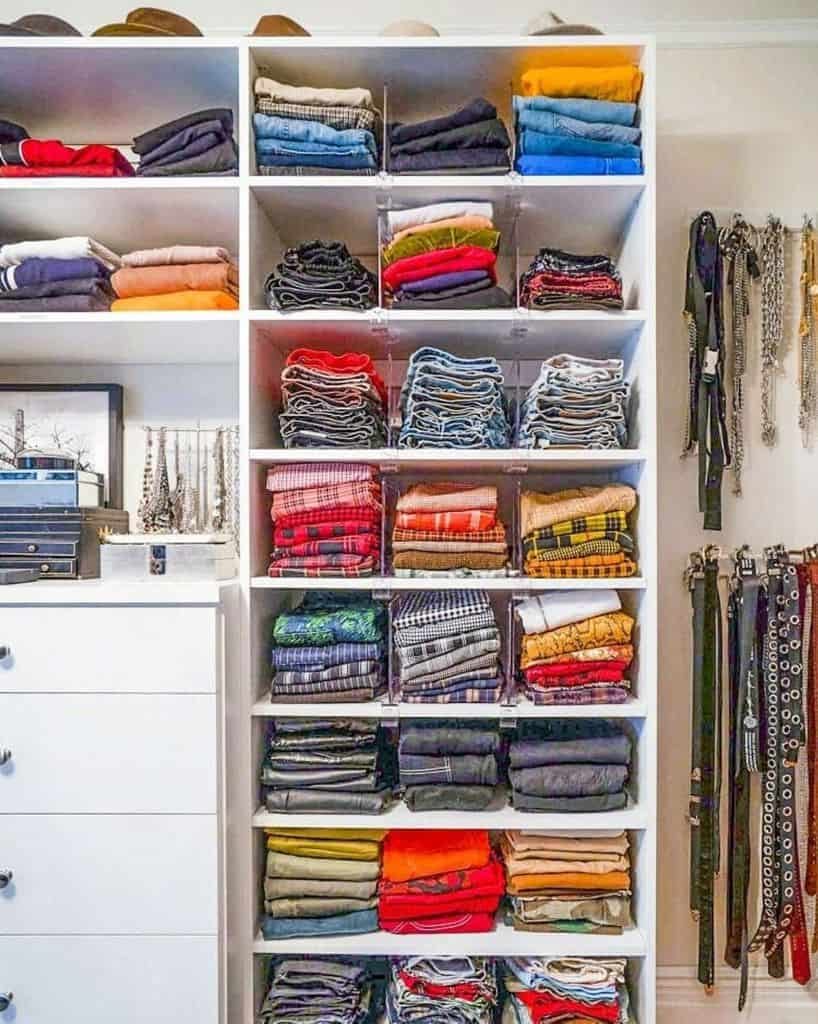 A neatly organized wardrobe with folded clothes sorted by color on shelves, belts and necklaces hang on the side