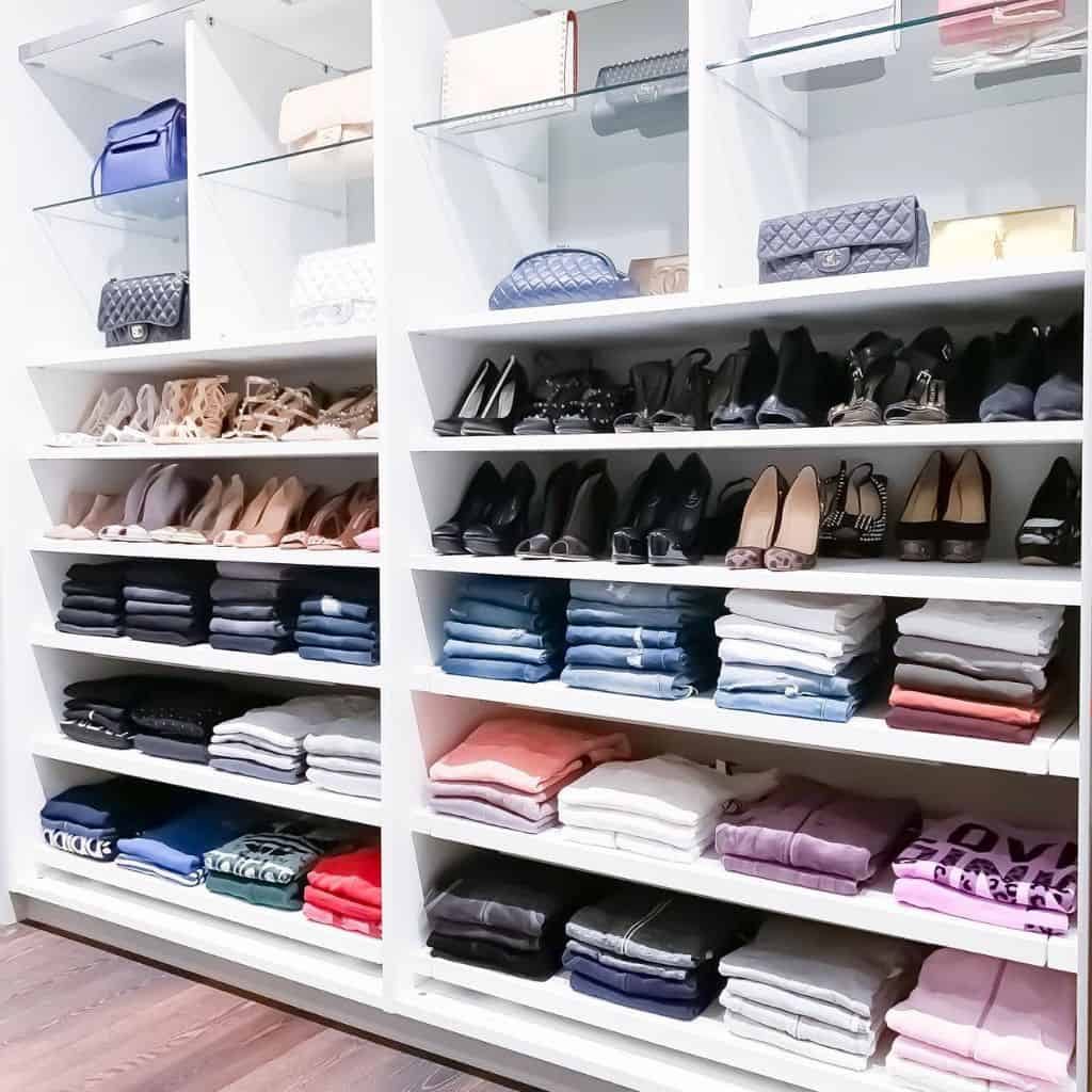 Organized closet with shelves of folded clothes, handbags, and pairs of shoes neatly arranged in a brightly lit space