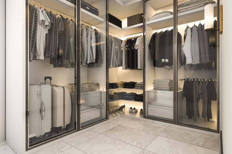 Modern walk-in closet with glass doors, neatly arranged clothes, folded items, luggage, and shoes on shelves and hangers