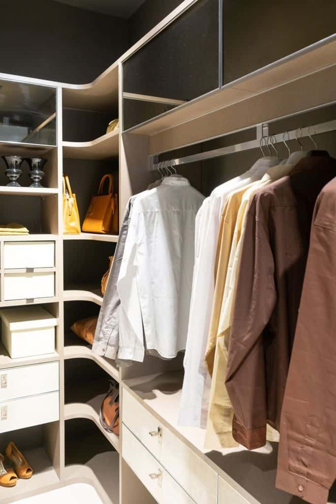 A walk-in closet with neatly hung shirts, shelves holding bags and shoes, and drawers for storage, all in a neutral color palette