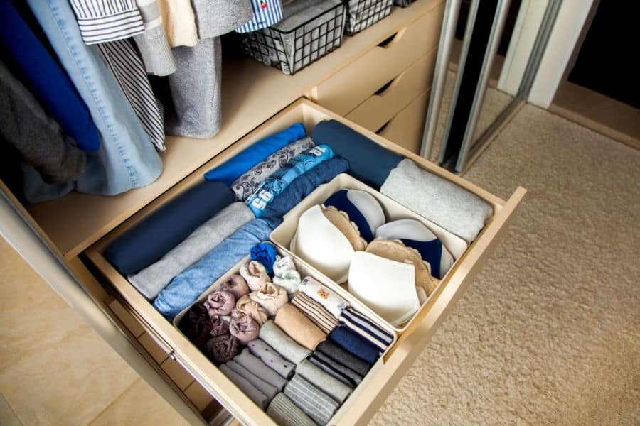 Organized drawer with neatly folded clothes, socks, and bras, next to a wardrobe with hanging clothes, beige carpet on the floor