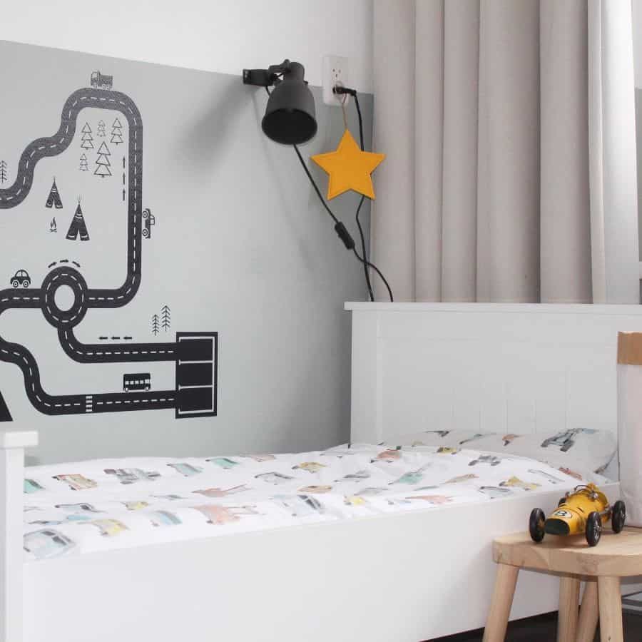 Minimalist kids' bedroom with a white bed, road map wall decal, black wall lamp, yellow star accent, and a toy car on a wooden stool