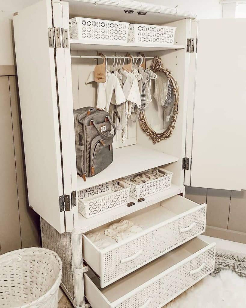 White wardrobe with open doors, hanging baby clothes, a gray backpack, a decorative mirror, and wicker storage baskets in drawers