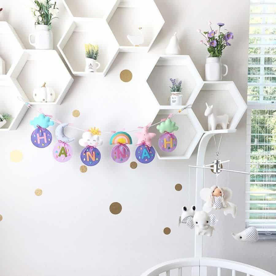 Bright and whimsical nursery with hexagon wall shelves, pastel name garland, gold polka dot accents, and a soft baby mobile