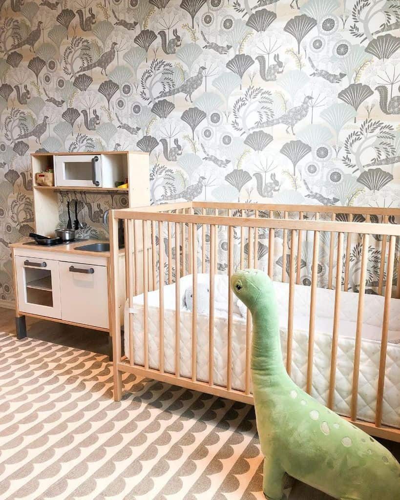 Nature-themed nursery with a wooden crib, botanical wallpaper featuring peacocks and foliage, a small play kitchen, and a large plush dinosaur toy on a patterned rug