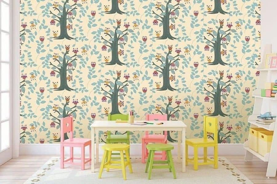 Colorful playroom with an owl and tree-themed wallpaper, a small white table surrounded by pink, green, and yellow chairs, and a white shelf with books and storage bins