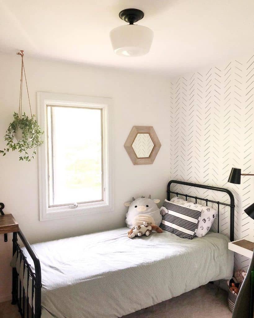 Minimalist kids' bedroom with a black metal bed frame, white and black accent pillows, a light-colored bedsheet, a geometric-patterned accent wall, a small wooden side table, a hanging plant, a hexagonal mirror, and a window allowing natural light