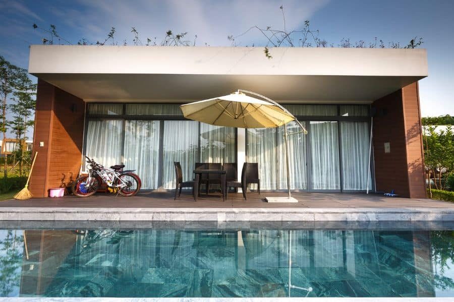 Modern pool house with floor-to-ceiling glass doors, a sleek patio, and a shaded dining area, offering a stylish and relaxing outdoor space