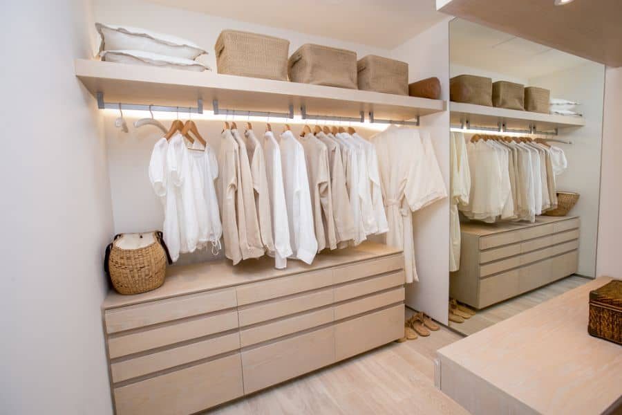 Minimalist closet with neatly hung beige and white clothes, wicker baskets on shelves, and drawers below in neutral tones throughout