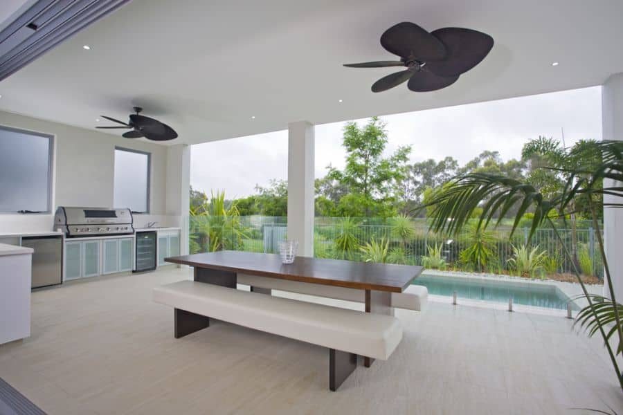 Modern outdoor kitchen and dining space with a built-in grill, sleek wooden table, ceiling fans, and a view of the pool and lush greenery