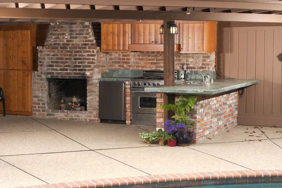 Rustic outdoor kitchen with exposed brick, a built-in fireplace, wooden cabinets, and a spacious bar area, perfect for poolside entertaining