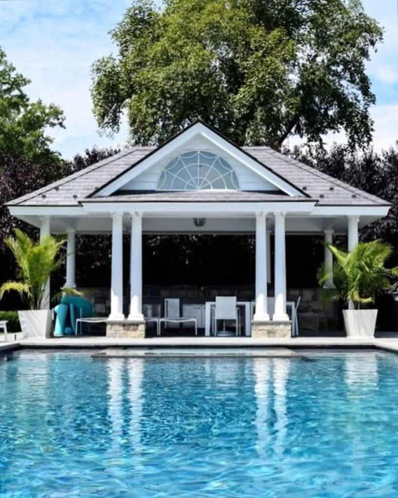 Elegant pool house with classic white columns, a gabled roof, and an outdoor dining area, overlooking a sparkling blue swimming pool