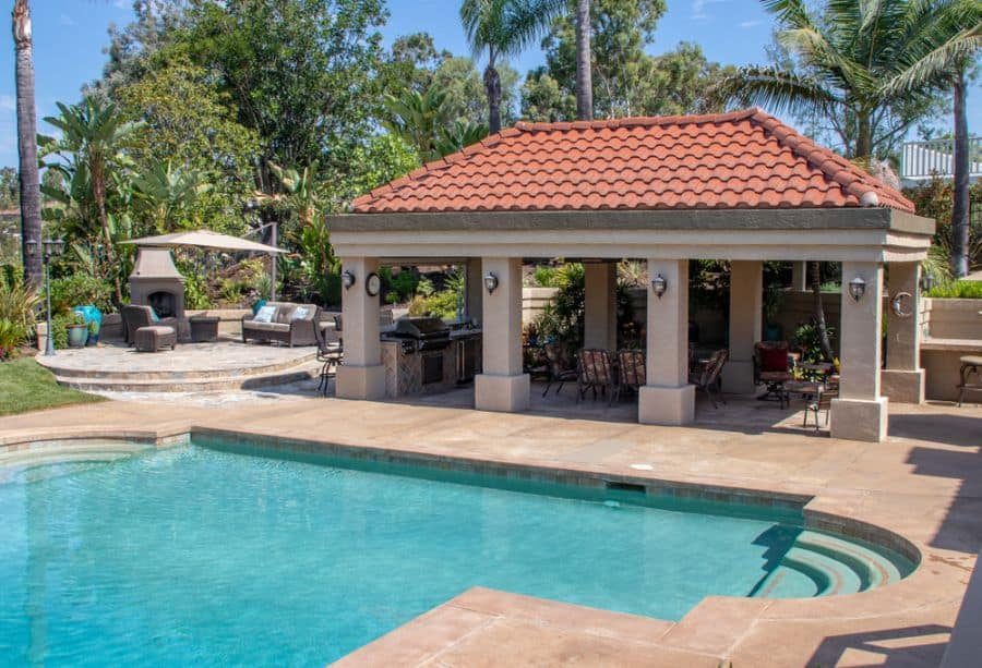 Luxury Mediterranean-style pool house with a red tile roof, outdoor kitchen, and cozy lounge area, surrounded by lush greenery and a sparkling pool