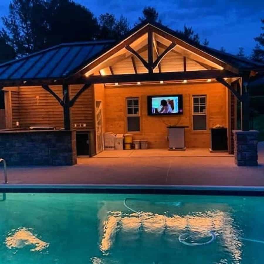 Cozy poolside pavilion with a rustic wooden design, warm lighting, an outdoor bar, and a TV, creating the perfect nighttime retreat