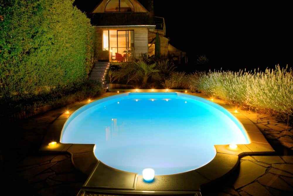 Elegant pool house with warm interior lighting, a glowing night-lit pool, and ambient garden lights creating a serene outdoor escape