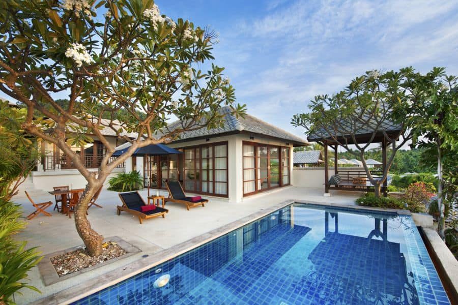 Luxury tropical pool house with a blue-tiled pool, sun loungers, and a shaded gazebo, surrounded by lush greenery and flowering trees