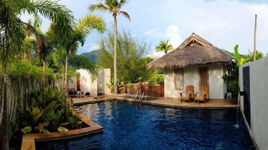 Tropical pool house with a thatched roof, wooden deck, and lush greenery, featuring a serene blue pool surrounded by palm trees and mountain views