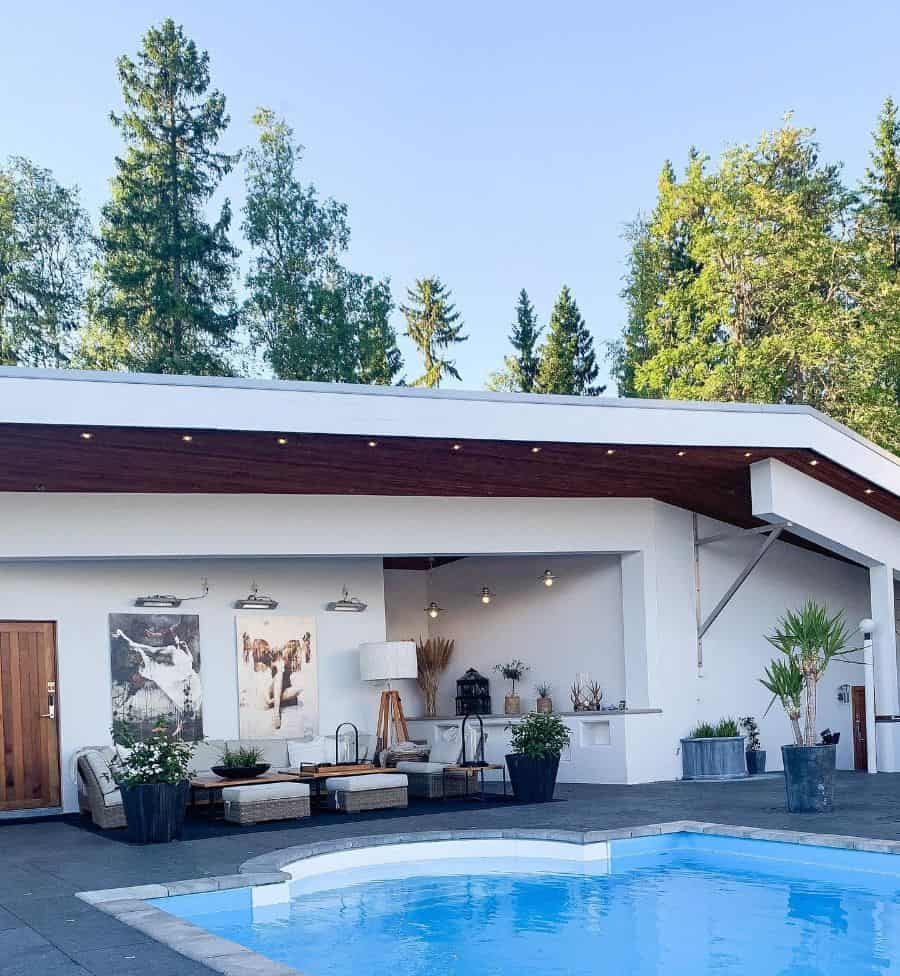 Modern pool house with a sleek white design, wooden accents, stylish outdoor seating, and a refreshing pool, surrounded by lush greenery