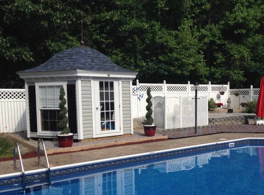 Charming poolside shed with a gray shingled roof, white siding, and glass-paneled doors, surrounded by greenery and a sparkling blue pool