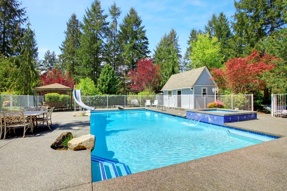 Spacious backyard pool area with a small pool house, waterslide, hot tub, and outdoor dining space, surrounded by lush trees and greenery