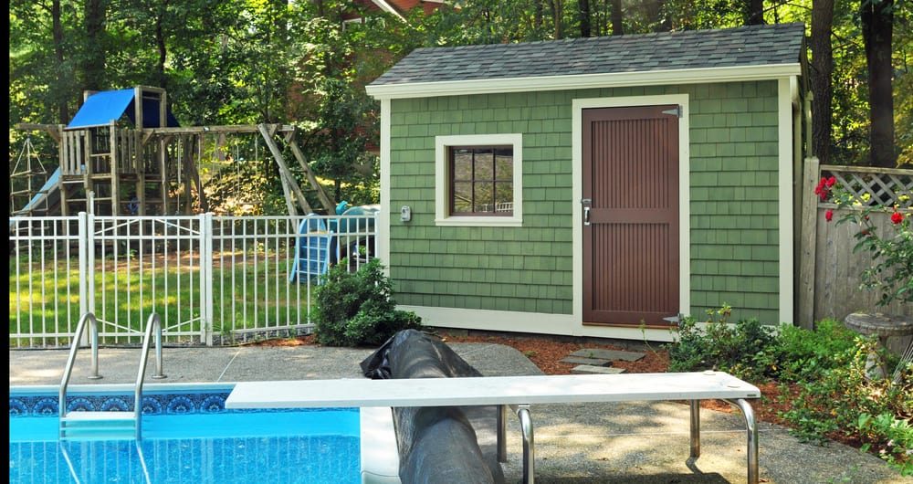 Charming green pool shed with shingle siding, a brown door, and a nearby diving board, set in a backyard with a playset and lush trees