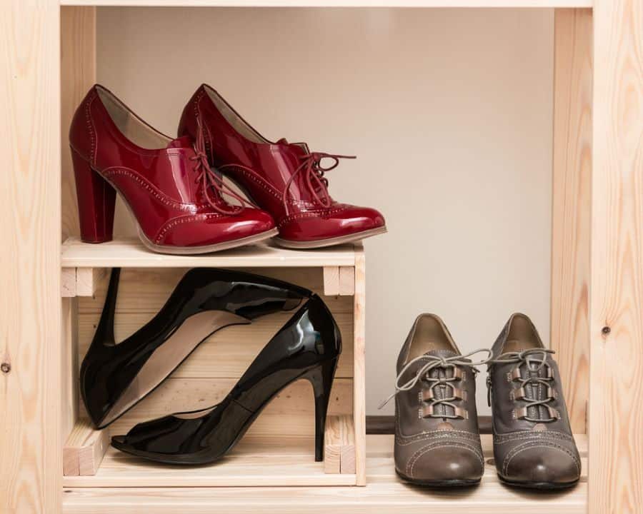 Three pairs of shoes on wooden shelves: red high-heeled shoes, black stilettos, and gray lace-up shoes