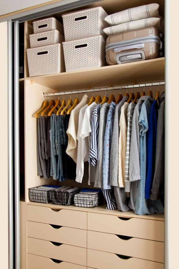 A neatly organized closet with baskets, folded linens, a suitcase, and clothes on hangers above drawers filled with wire baskets