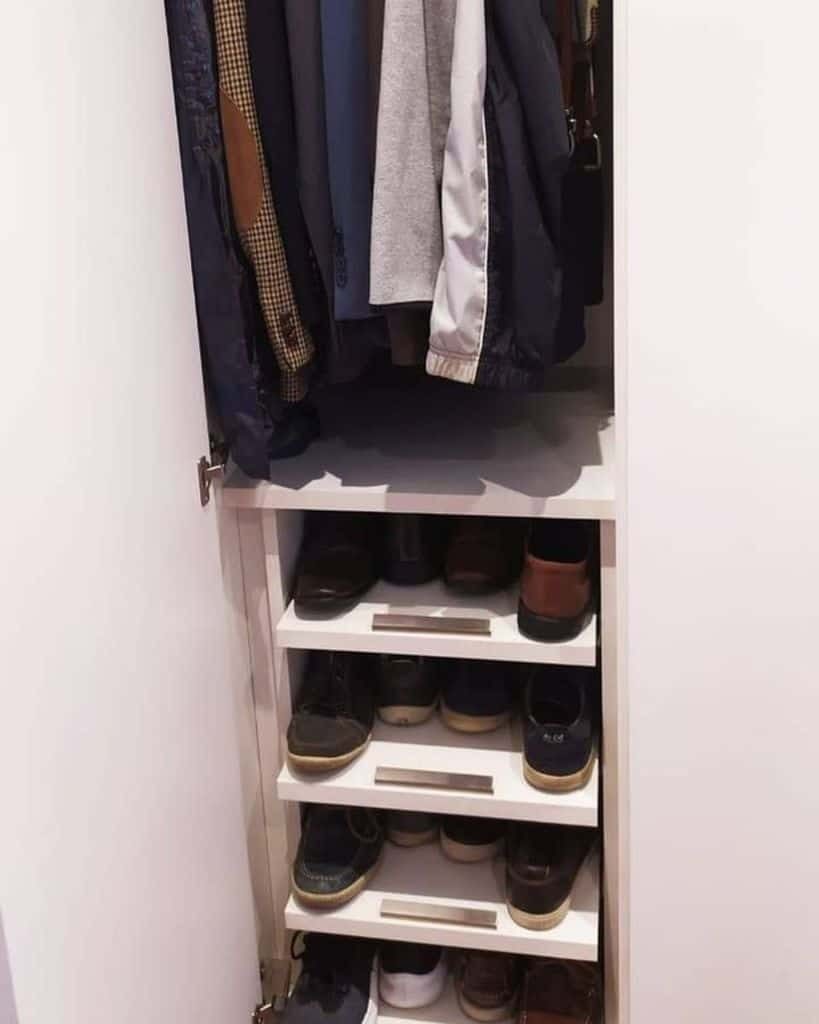 A small closet with jackets and shirts hanging above three shelves filled with various pairs of shoes