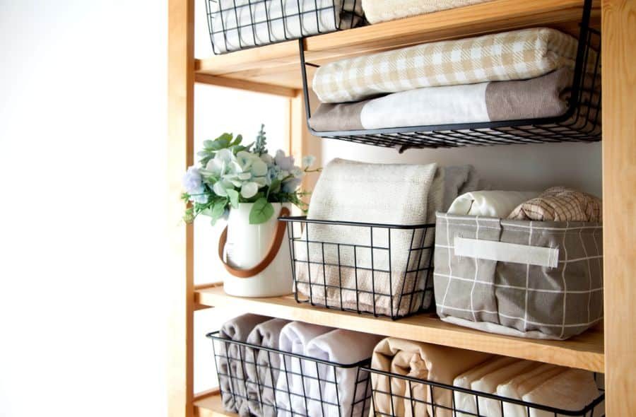 Wooden shelving with wire baskets holding folded blankets and linens, with a white pitcher containing flowers also on the shelf