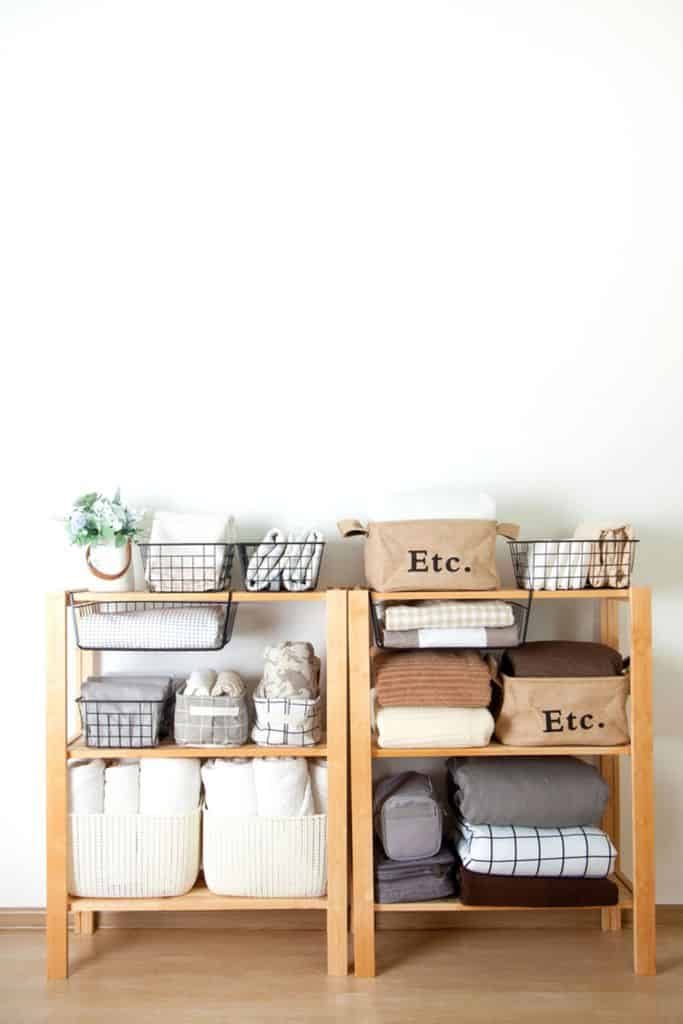 Wooden shelving unit with neatly organized baskets and folded linens, neutral colors, minimal decor, and spacious white wall backdrop