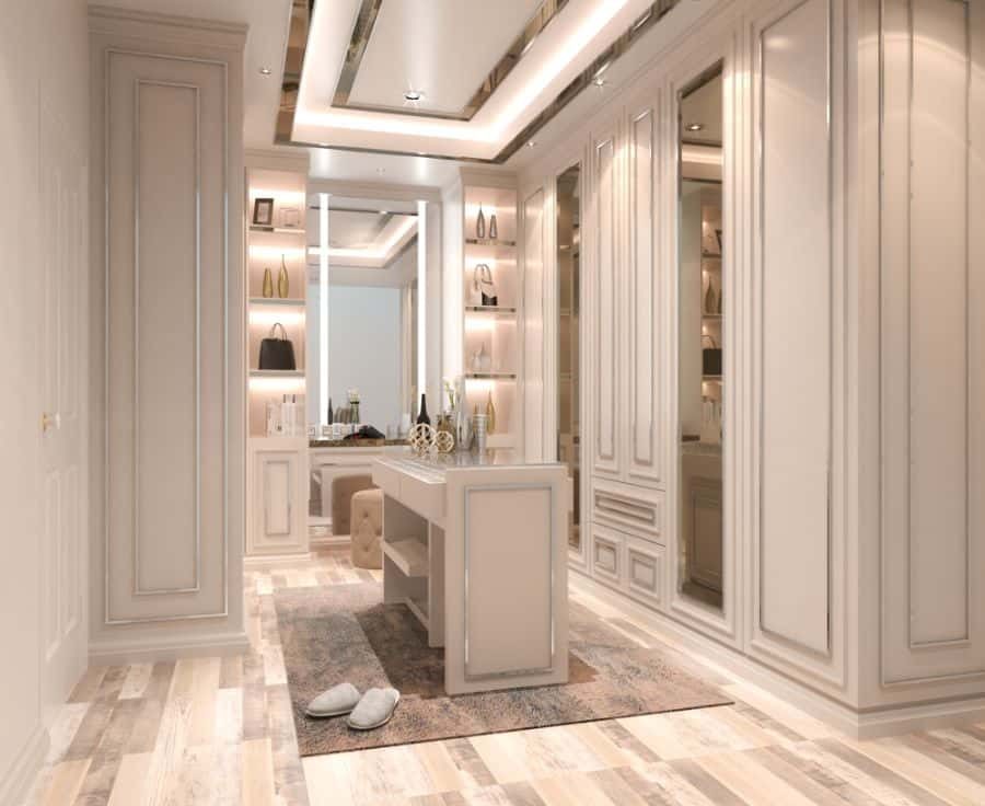 Luxurious beige walk-in closet with a mirror, shelves displaying bags, a central island, and slippers on a rug on wooden floor