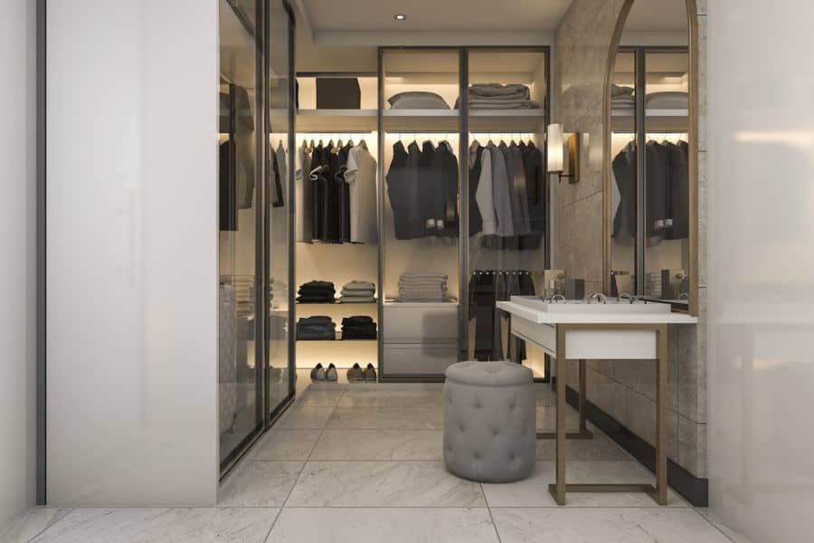 Modern walk-in closet with glass doors, shelves neatly stacked with clothes, and a small vanity with a cushioned stool in front