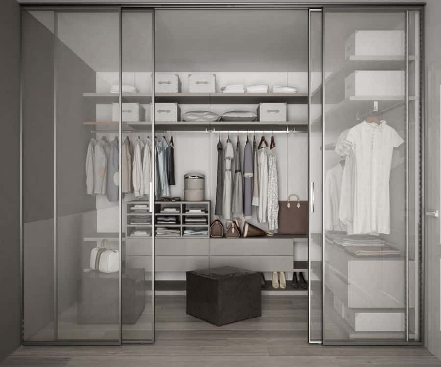 Modern closet with glass sliding doors featuring neatly organized clothes, boxes, and bags on shelves and a rack, with a dark ottoman below