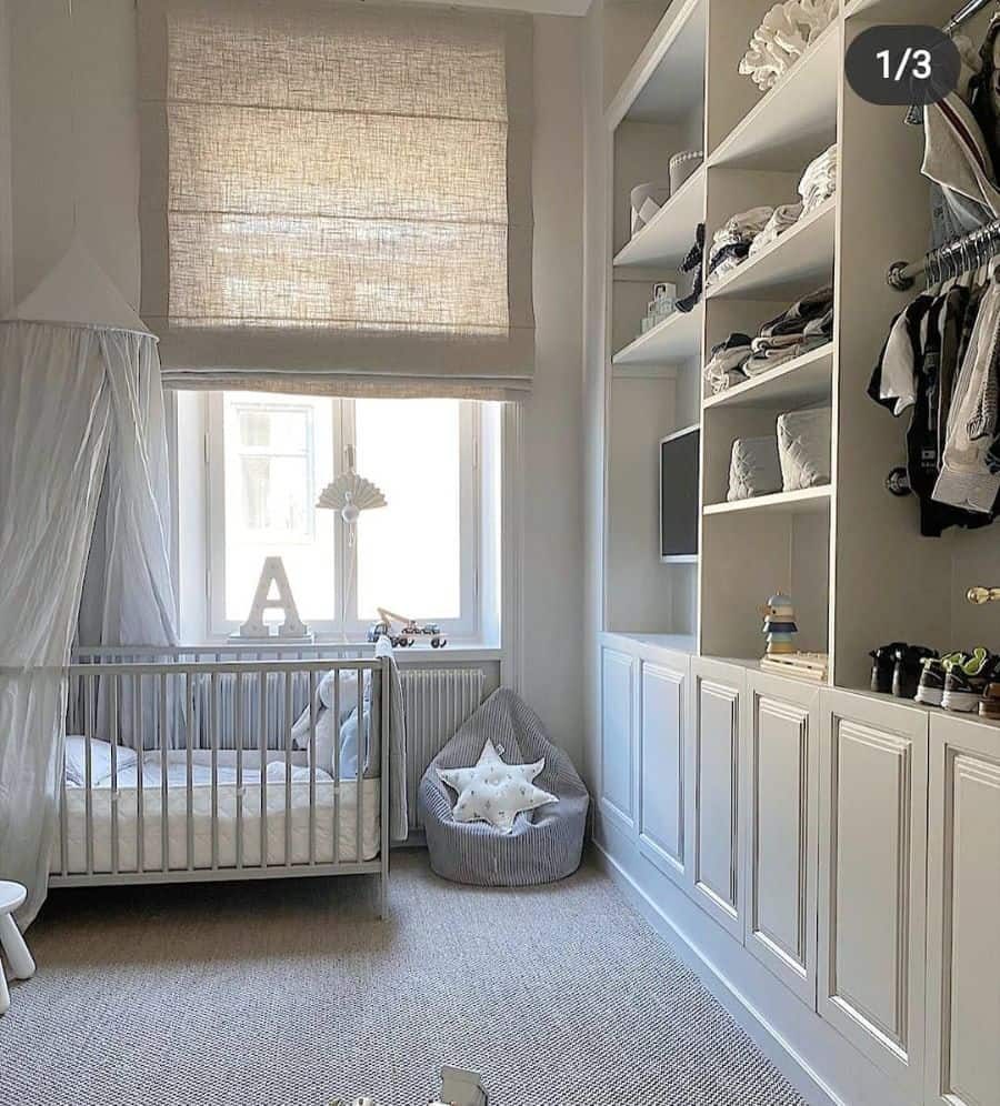 Minimalist white nursery featuring a crib with a sheer canopy, a built-in wardrobe with open shelving, a cozy bean bag with a star pillow, and a large window with beige Roman shades allowing natural light to brighten the space