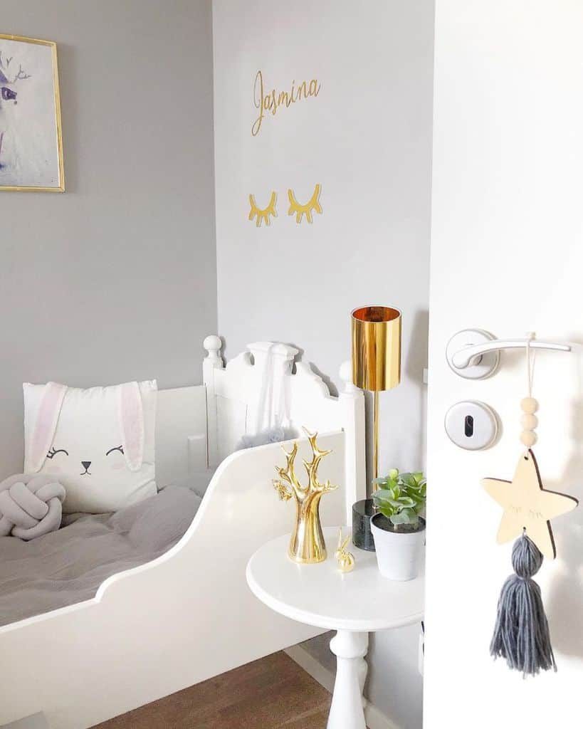 Charming white kids' bedroom with a vintage-style bed featuring a bunny-themed pillow, a cozy gray blanket, and a small round bedside table adorned with gold decor, potted plants, and a sleek gold lamp