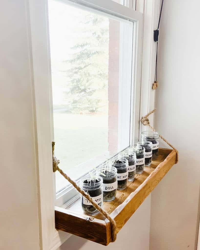 Jars with plants labeled on a wooden hanging shelf by a window, suspended with ropes