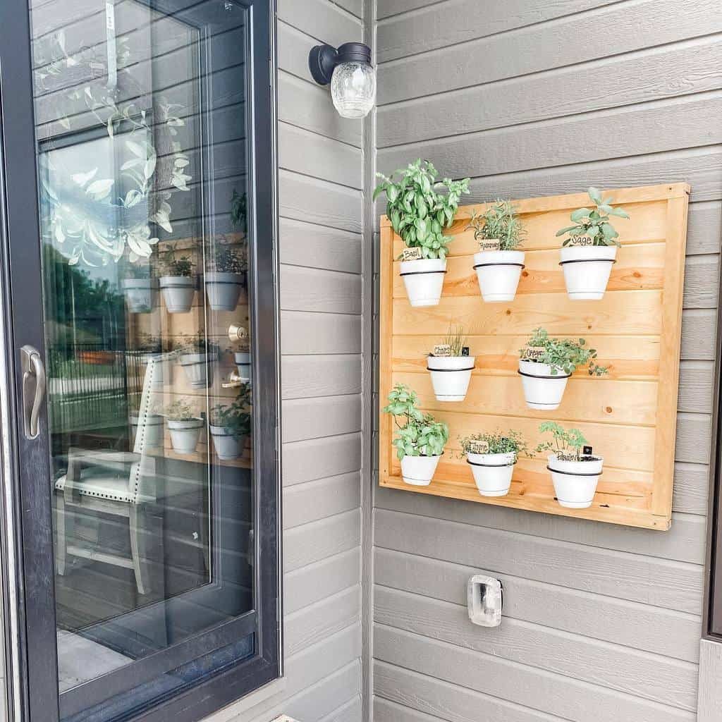 Vertical herb garden with nine small pots on a wooden board, mounted on a gray wall next to a window 